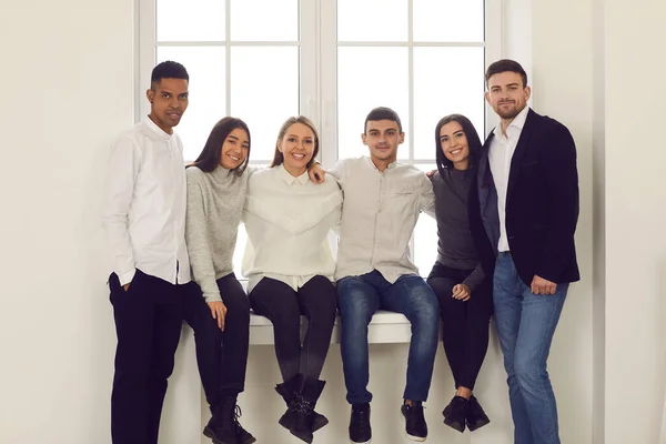 Группа молодых улыбающихся людей разных национальностей, сидящих на подоконнике и обнимающихся — стоковое фото