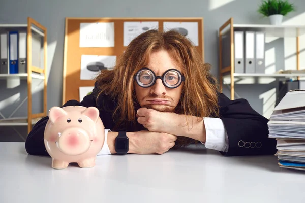 Ofis masasında domuz kumbarasıyla oturup para kazanmanın yollarını düşünen komik bir adam. — Stok fotoğraf