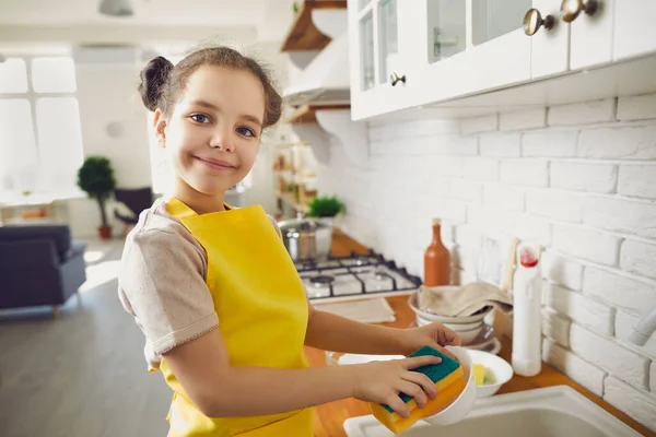 Portrait of a cute little girl in a bright apron washing dishes in a bright kitchen.