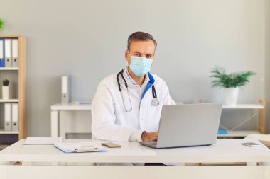 Masasında dizüstü bilgisayarla oturmuş kameraya bakan koruyucu maskeli ciddi bir doktor..