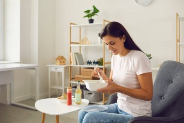 Mutlu genç bir kadın sağlıklı bir yemek yiyor. Evdeki koltukta oturuyor.