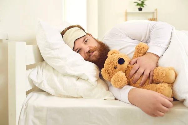 Pria dewasa berbaring tanpa tidur di tempat tidur yang lembut, memeluk boneka beruang dan melihat kamera — Stok Foto