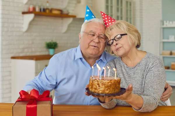 Felice coppia matura in cappelli cono soffiando candele sulla torta che celebra il compleanno a casa. — Foto Stock