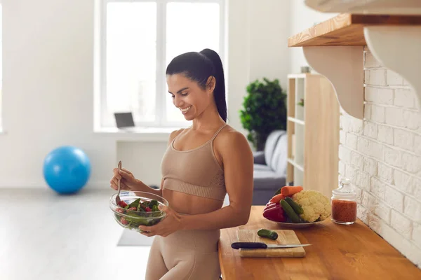 Mulher fitness esbelta em sportswear de pé em casa na cozinha com uma tigela de salada fresca. — Fotografia de Stock