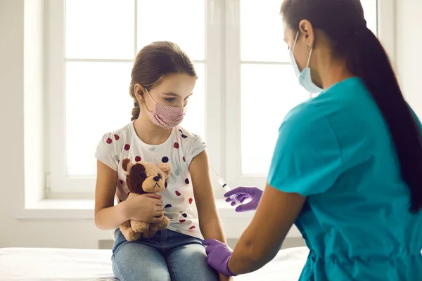 Arzt injiziert Impfstoff in die Hand eines süßen kleinen Mädchens, das ihr Lieblingsspielzeug hält. — Stockfoto