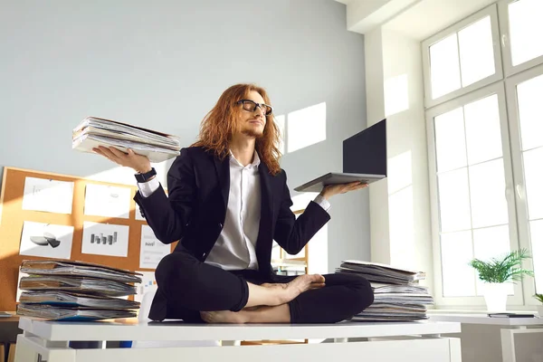 Calm worker holding papers and laptop and meditating in yoga pose on office desk
