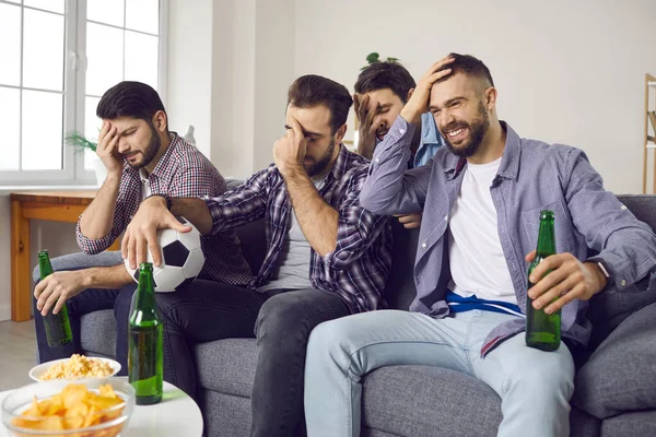 Disappointed fans sitting on sofa at home and watching terrible soccer game on TV