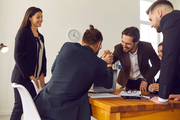 Dos jóvenes empresarios felices estrechando la mano en la oficina con compañeros de trabajo sonrientes mirando —  Fotos de Stock