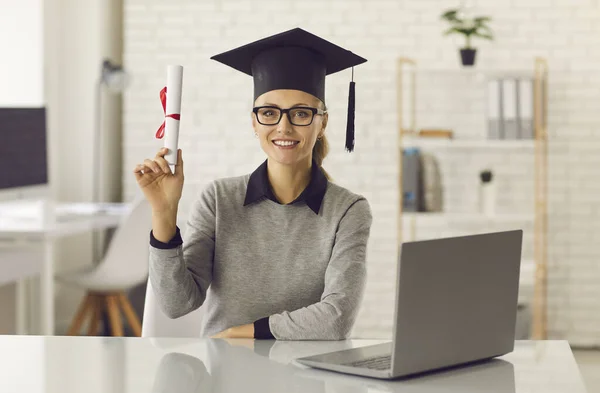 Talangfulla online kurser examen sitter vid skrivbordet med bärbar dator och hålla hennes diplom — Stockfoto