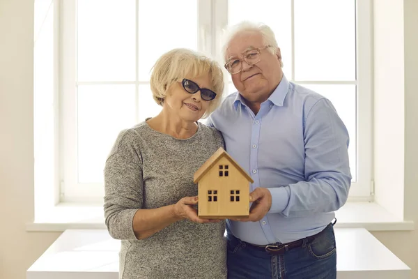 Felice coppia anziana matura in piedi insieme, abbracciando e tenendo piccola casa decorativa in legno — Foto Stock