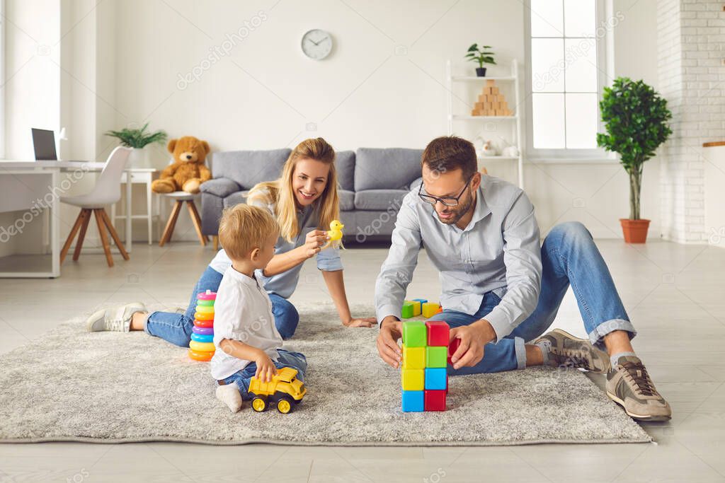 Mom and dad play with their son at home with a variety of toys and colored cubes sitting in a room.