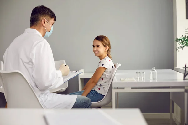 Lekarz pediatra w mundurze medycznym i masce rozmawia z małą uśmiechniętą pacjentką — Zdjęcie stockowe