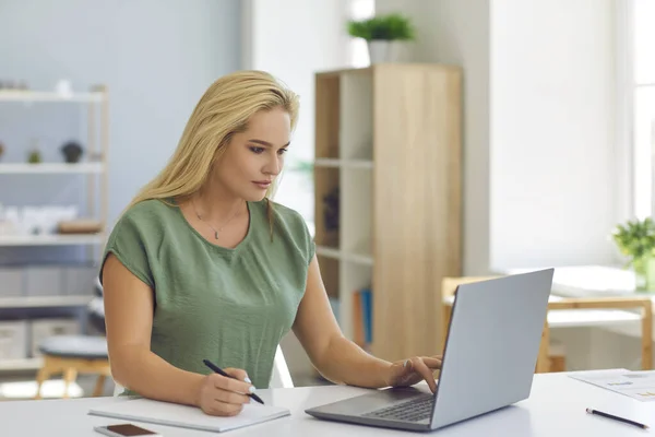 Frau sitzt mit Laptop am Schreibtisch, schaut Lerntutorial oder Online-Vorlesung und macht sich Notizen — Stockfoto