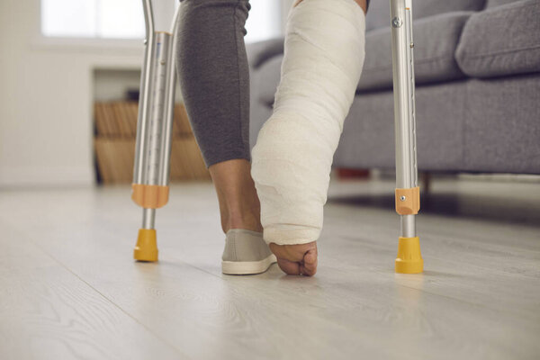 Close up of a womans leg with a broken leg in a cast walking with crutches in the living room.