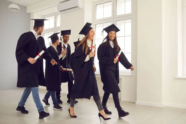 Multiracionális egyetemi diplomások csoportja sétál végig a folyosón diplomával a kezében. — Stock Fotó