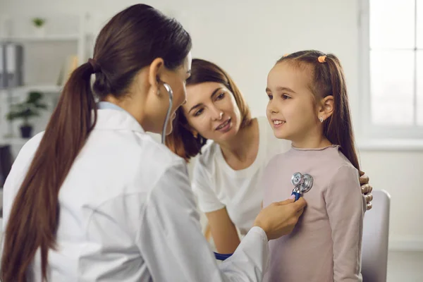 Молодая женщина врач-педиатр в медицинской форме осматривает маленькую улыбающуюся девочку со стетоскопом — стоковое фото