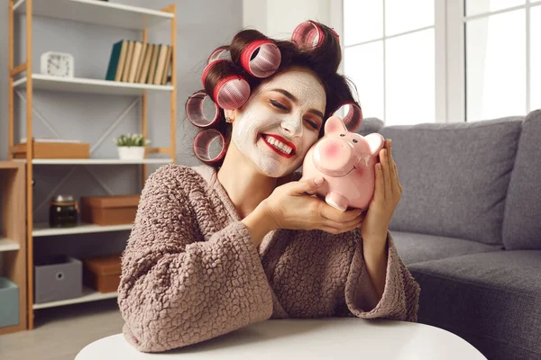Happy woman with face mask and hair curlers hugging piggy bank, smiling and dreaming