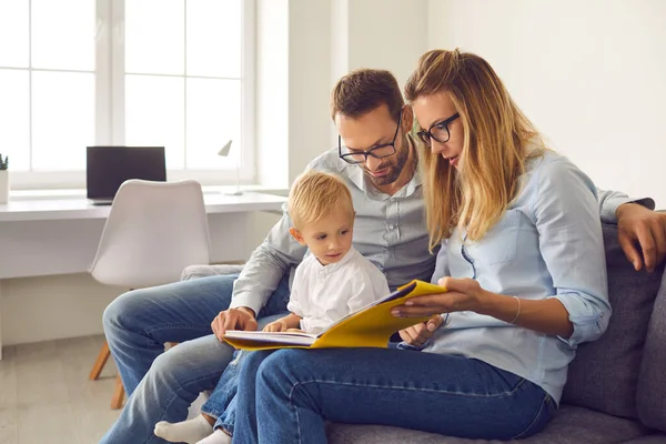 Mamma, papà e figlio leggono un libro di storie insieme seduti sul divano in una stanza luminosa a casa. — Foto Stock