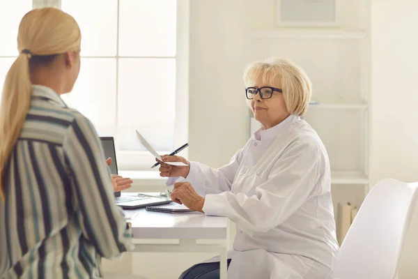 Freundliche Oberärztin gibt einer Patientin ein Rezept, während sie an einem Tisch in der Klinik-Praxis sitzt. — Stockfoto