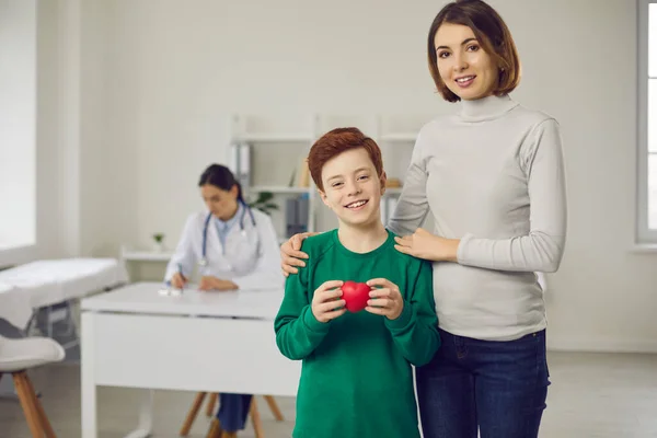 Uśmiechnięty chłopiec i jego matka stojący z czerwonym sercem po wizycie u lekarza pediatry — Zdjęcie stockowe
