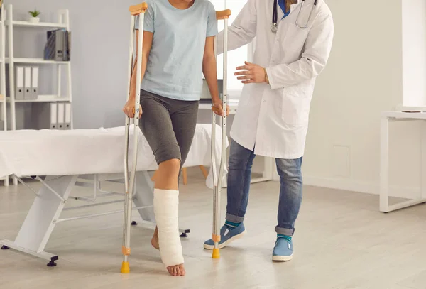 Paciente com perna quebrada caminhando com muletas assistidas pelo médico durante visita ao hospital — Fotografia de Stock