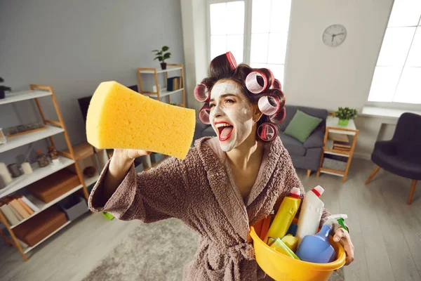 Divertido loco joven ama de casa en rulos y cuidado de la piel mascarilla ordenando su casa — Foto de Stock