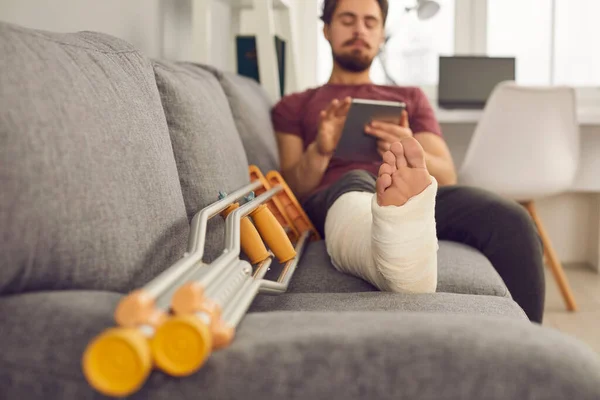 Jovem com a perna partida no molde sentado no sofá com muletas e usando tablet — Fotografia de Stock