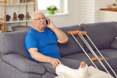 Kırık bacaklı yaşlı adam kanepede oturuyor ve telefonda doktor ya da aileyle konuşuyor.