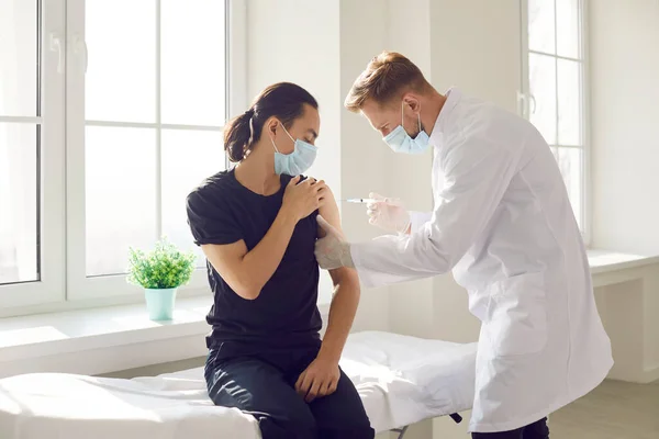 Manlig läkare i medicinsk mask kommer att injicera ett vaccin i en ung patients hand.. — Stockfoto