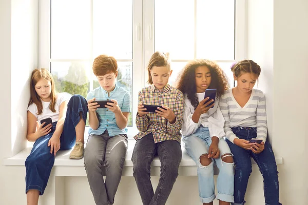 Crianças sentadas no peitoril da janela, jogando jogos online em telefones celulares e ignorando uns aos outros — Fotografia de Stock