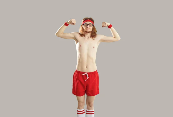 Funny hipster skinny young guy in red shorts and naked torso standing and showing biceps