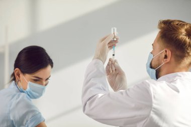 Üniformalı, maskeli ve eldivenli bir adam aşıdan önce şırınga dolduruyor.