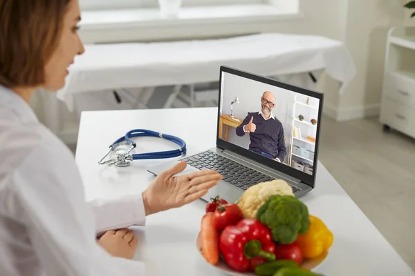 Online doctor sitting at desk with laptop and video calling happy senior patient