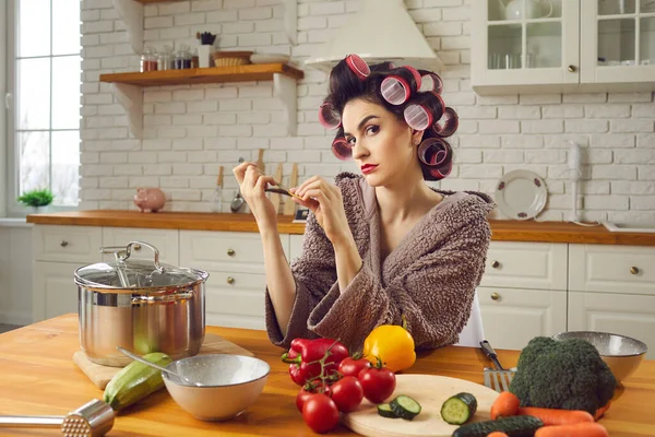 Jovem irritada em encrespadores de cabelo sentado na mesa da cozinha, arquivando unhas e pensando — Fotografia de Stock