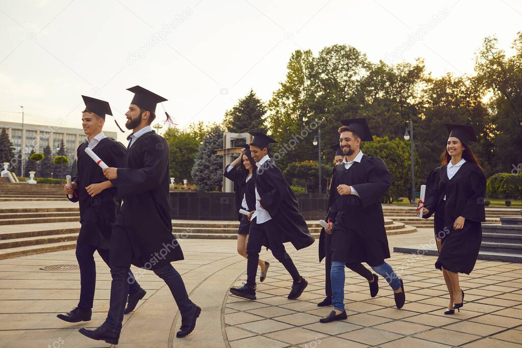 Group of young happy university graduates in traditional mantles walking with diplomas