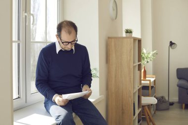 Evdeki pencere eşiğinde otururken bazı mektuplar ve belgeler okuyor.