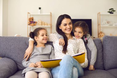 Gülümseyen anne ve küçük kızları kanepede oturmuş kitap okuyor, sarılıyor ve yan bakıyor.