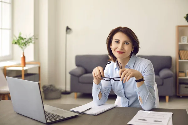 Happy businesswoman sitting at office desk with laptop, smiling and looking at camera