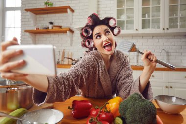 Saç bigudili kadın mutfakta yemek pişirirken cep telefonundan komik selfie çekiyor.