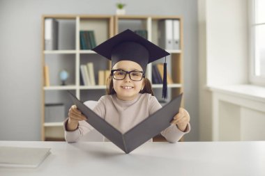 Gözlüklü küçük kız elinde diplomasıyla mezuniyet şapkasıyla masada oturuyor..