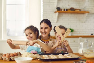 Mutlu genç anne ve kızları fırın sırasında merdane ve ahşap spatulalar kullanıyorlar.