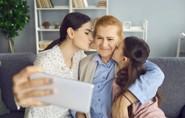 Happy daughter and granddaughter kissing grandma and taking selfie on smartphone