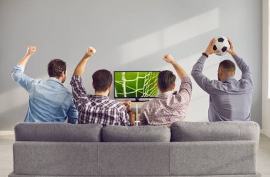 Kolları havada dört yetişkin erkek arkadaş evde televizyonda futbol maçı izliyorlar..