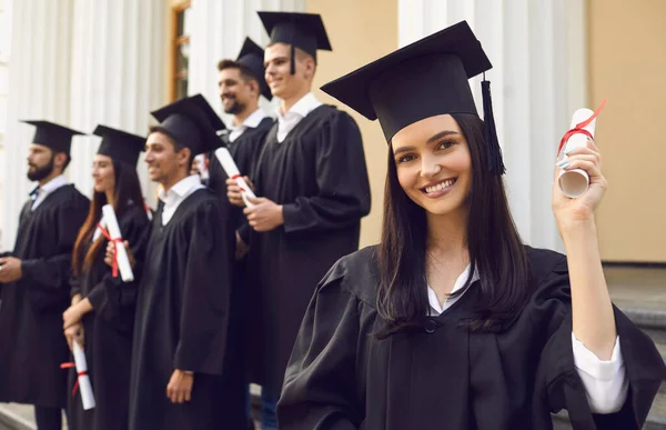 Young smiling girl university graduate standing holding diploma in raised hand over group of mates
