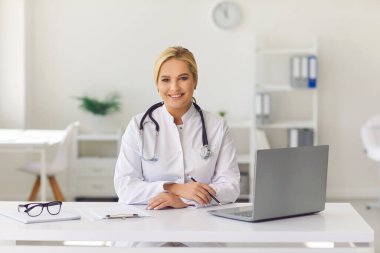 Dost canlısı genç bir kadın, doktor olarak çalışıyor, hastane ofisinde masasında oturuyor, gülümsüyor ve kameraya bakıyor..