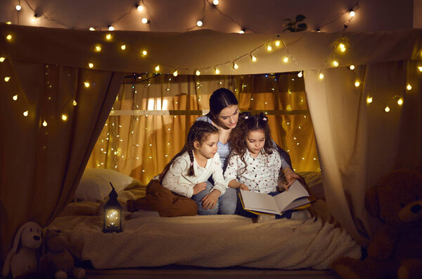 Mom and kids reading book of fairy tales sitting in blanket fort in cozy dark playroom
