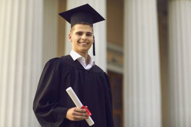 Mezun olmuş genç bir çocuk, elinde diplomasıyla üniversite binasının arka planında duruyor.
