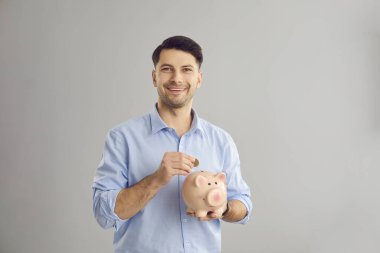 Mutlu sakallı adam kumbaraya para koyuyor ve gri kameraya bakıyor.