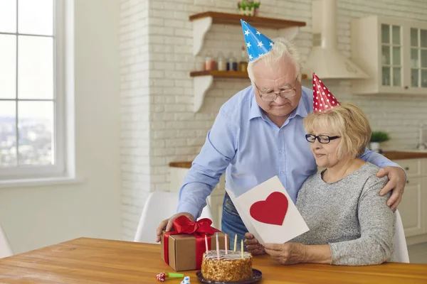 Senior husband congratulating his wife on her birthday, giving her cake, present and greeting card