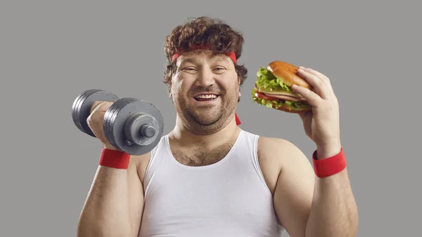 Смішний пухкий чоловік тримає гантелі, їсть великий смачний бургер і посміхається на камеру — стокове фото